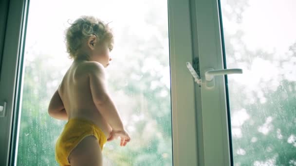 Маленька дівчинка в жовтих трусиках дивиться дощ біля вікна вдома — стокове відео
