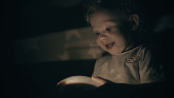 Bebê pequeno feliz olha para brinquedo projetando luminoso no quarto escuro — Vídeo de Stock