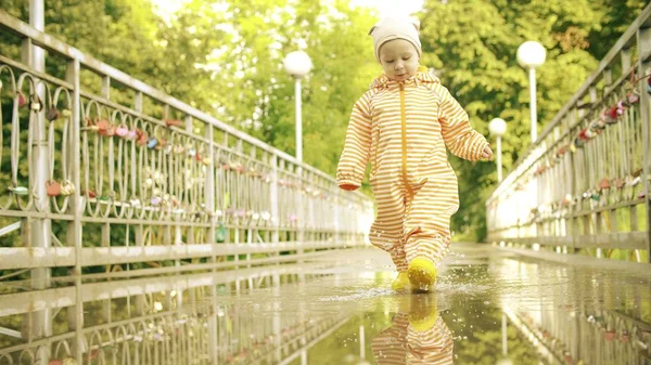 Забавная маленькая девочка в оранжевом водонепроницаемом плаще и резиновых ботинках дождя ходит по луже после дождя — стоковое фото