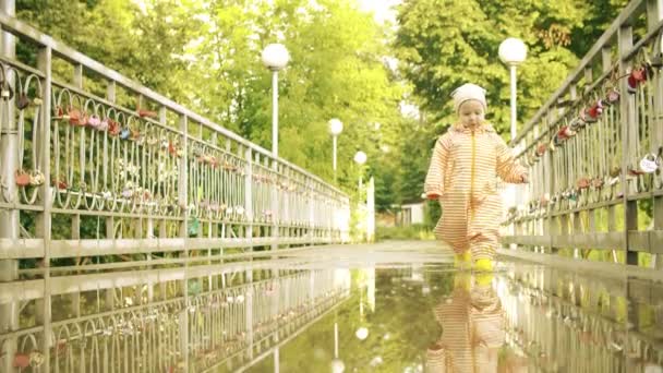 Menina engraçada em laranja impermeável capa de chuva e botas de chuva de borracha caminha na poça após a chuva, câmera lenta — Vídeo de Stock