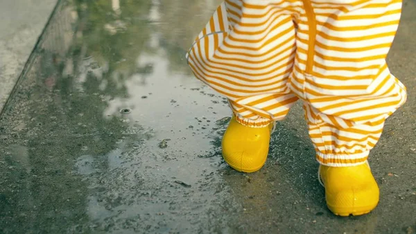Pequeño bebé con impermeable a rayas naranja y botas de lluvia de goma amarilla camina sobre charcos después de la lluvia, primer plano —  Fotos de Stock