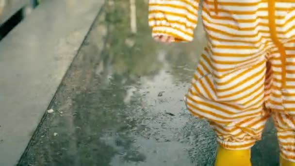 Bebê pequeno em capa impermeável listrada laranja e botas de chuva de borracha amarela caminha em poças após a chuva, close-up — Vídeo de Stock