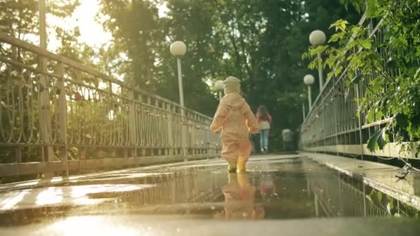 Bambina in impermeabile arancione impermeabile impermeabile e stivali da pioggia in gomma cammina su pozzanghere a sua madre, scatto al rallentatore — Video Stock