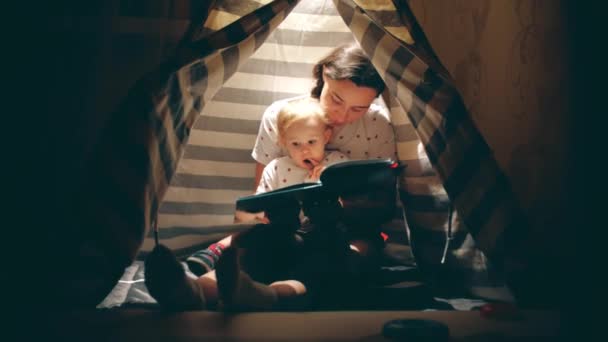 Mãe e seu bebê pequeno leram um livro juntos em um chá iluminado aconchegante à noite — Vídeo de Stock