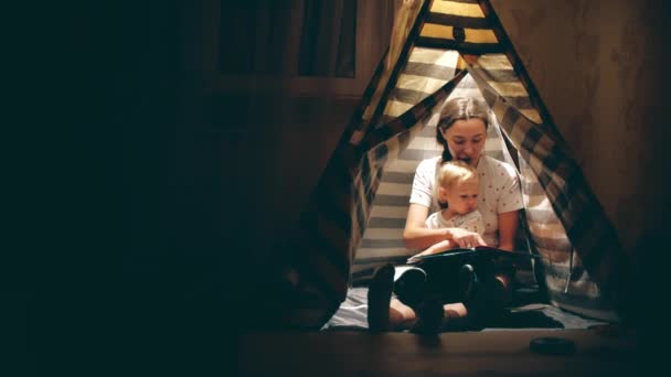 Mama en haar kleine dochter lezen een boek samen in een tipi in de avond — Stockvideo