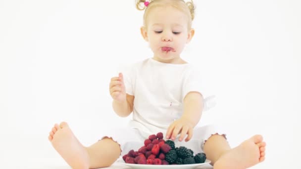 Linda niña rubia come bayas contra fondo blanco — Vídeos de Stock