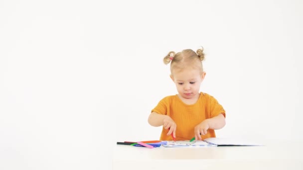 Little blonde baby girl coloring picture with two colorful crayons against white background — Stock Video