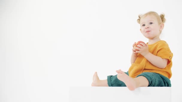 Baby met oranje en groene kleren eet Apple tegen witte achtergrond — Stockvideo