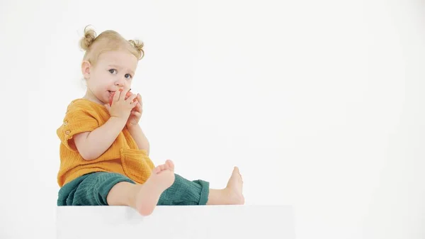 Nachdenkliches Baby in orangefarbener und grüner Kleidung, das Apfel auf weißem Hintergrund isst — Stockfoto