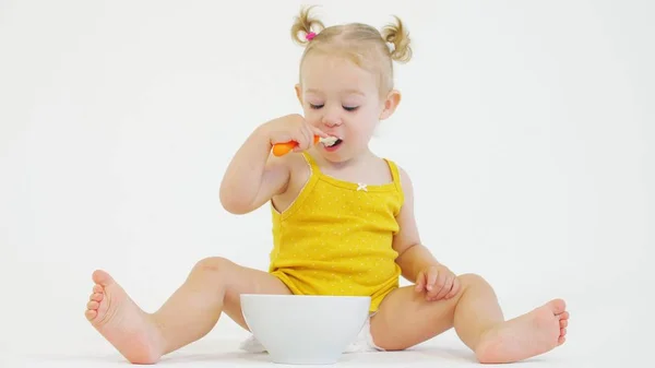 Menina bebê comendo seu café da manhã com uma colher no fundo branco — Fotografia de Stock