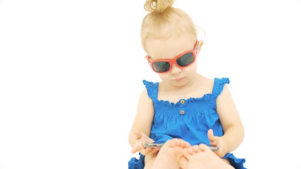 Πορτρέτο ενός μωρού κορίτσι που φοράει γυαλιά ηλίου και μπλε φόρεμα με ένα μοντέρνο smartphone. Εφαρμογές που σχετίζονται με τις διακοπές — Αρχείο Βίντεο