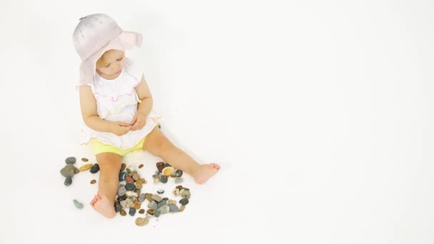 Liten blond flicka som bär hink hatt leker med havet sten och skal på vit bakgrund — Stockvideo