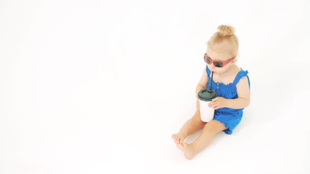 Baby meisje dragen zonnebril drankjes uit papieren beker met een stro op witte achtergrond — Stockvideo