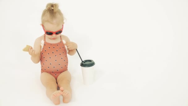 Μωρό κορίτσι φορώντας γυαλιά ηλίου και καφέ μαγιό που έχουν ένα σνακ σε λευκό φόντο — Αρχείο Βίντεο