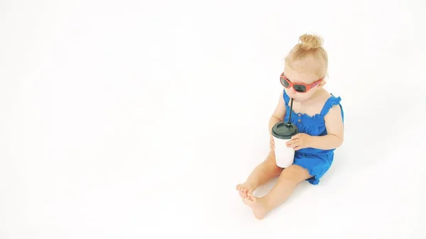 Bebê menina usando óculos de sol bebidas de copo de papel com uma palha no fundo branco — Fotografia de Stock