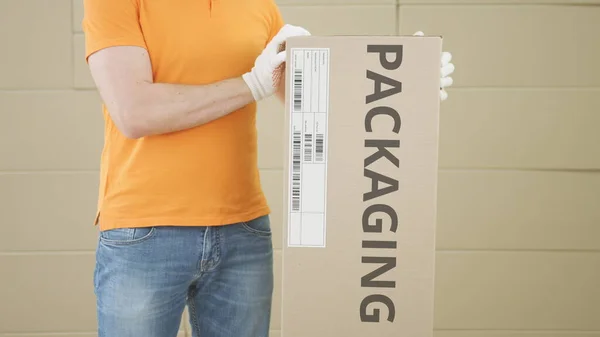 Homem carregando grande caixa de papelão com texto PACKAGING contra fundo pilhas de papelão — Fotografia de Stock