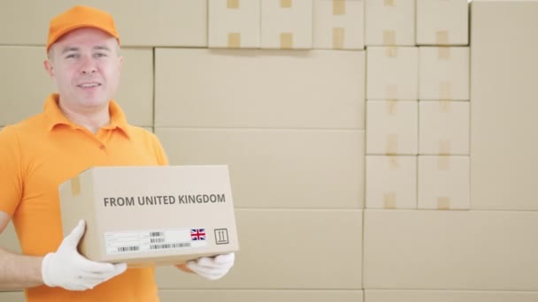 Pracownik noszący pomarańczowy mundur trzyma pudełko z nadrukiem OD ZJEDNOCZONEGO KRÓLESTWA tekst i etykieta — Wideo stockowe