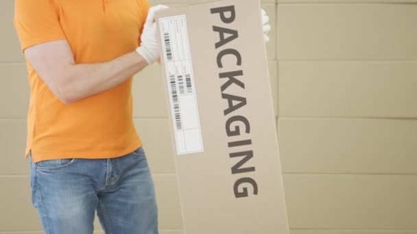 Ο άνθρωπος που μεταφέρει μεγάλο κουτί από χαρτόνι με το κείμενο ΣΥΣΚΕΥΑΣΙΑ σε φόντο στοίβες χαρτοκιβωτίων — Αρχείο Βίντεο