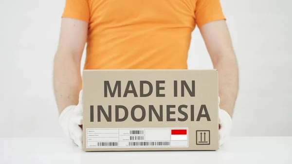 Trabajador vistiendo uniforme naranja pone caja con HECHO EN INDONESIA imprimir sobre la mesa —  Fotos de Stock
