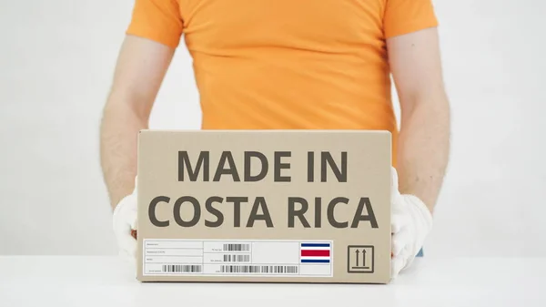 Trabajador vistiendo uniforme naranja pone la caja con la impresión MADE IN COSTA RICA en la mesa —  Fotos de Stock