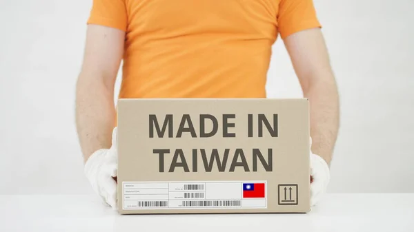 Trabajador vistiendo uniforme naranja pone caja con HECHO EN TAIWAN imprimir en la mesa —  Fotos de Stock