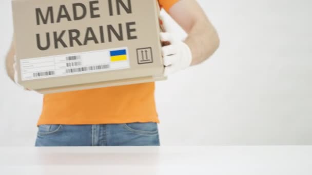 Pracovník v oranžové uniformě vloží krabici s potiskem MADE IN UKRAINE na stůl — Stock video