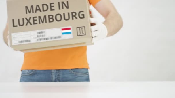 Pracovník v oranžové uniformě vloží krabici s potiskem MADE IN LUXEMBOURG na stůl — Stock video