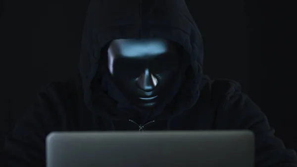 Hacker desconhecido vestindo roupas escuras e máscara preta funciona em seu laptop — Fotografia de Stock