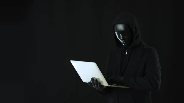 Hacker misterioso vestindo roupas escuras e máscara preta usa seu laptop — Fotografia de Stock