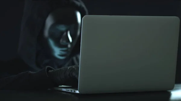 Un uomo sconosciuto che indossa una maschera nera ruba informazioni da un portatile. Nozioni di hacking o darknet — Foto Stock