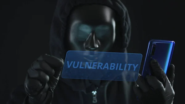 El hacker con máscara negra extrae la pestaña VULNERABILIDAD de un teléfono inteligente. Concepto de hackeo —  Fotos de Stock
