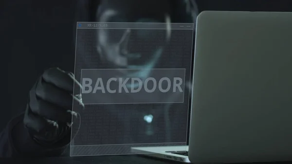 Un hacker con máscara negra saca la pestaña BACKDOOR de un portátil. Concepto de hackeo —  Fotos de Stock