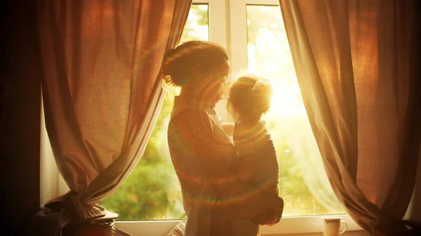 Mamma håller och kramar sin lilla dotter nära fönstret hemma, familjens tid — Stockfoto