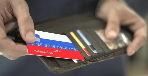 Мужчина вытаскивает пластиковую банковскую карту с флагом Словении из бумажника, вымышленный номер карты — стоковое фото