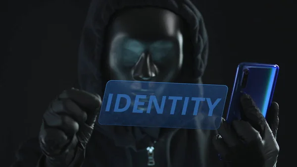 Hacker con máscara negra tira de la pestaña IDENTIDAD desde un teléfono inteligente —  Fotos de Stock