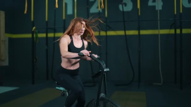 Rödhårig attraktiv fitness kvinna ridning motionscykel i gymmet. En smal dam på cykel. Kvinnlig idrottsman gör intensiv träning på gym cykel — Stockvideo