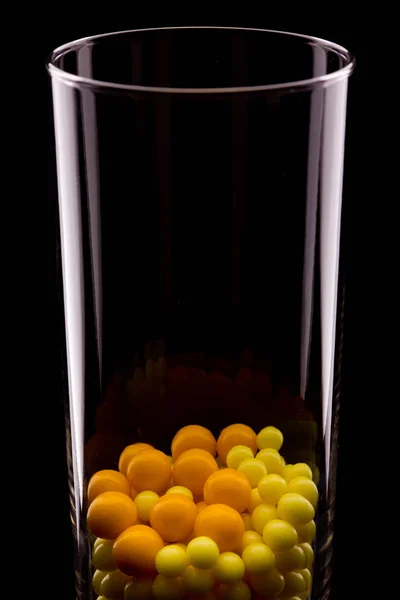 En un vidrio transparente hay comprimidos redondos de color amarillo y rojo. Un vaso se refleja en el negro. Fondo oscuro . — Foto de Stock