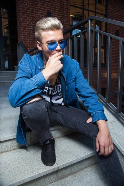 Guapo joven rubio en gafas de sol azules fumando en las escaleras — Foto de Stock