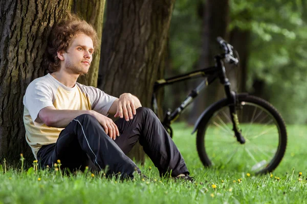 Young cyclist with fury hair sitting by a tree in a park on the grass — Stock Photo, Image