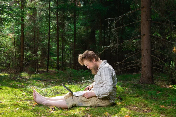 shot of confused and surprised Caucasian bearded hipster feeling frustrated, sitting at outdoor in the wood with generic laptop pc, trying to concentrate on work, looking exhausted.