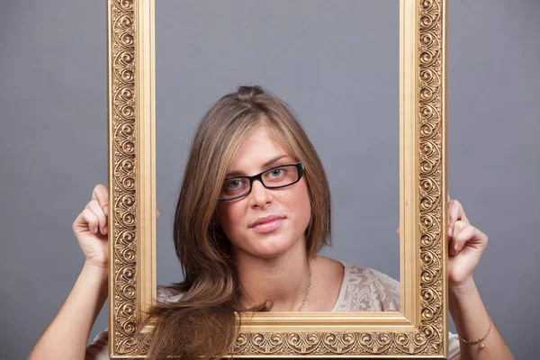 Young woman in eyeglasses looking through wooden golden frame — Stock Photo, Image