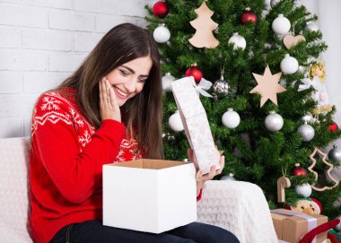 Genç, mutlu, şaşkın kadın süslü Noel ağacının yanındaki hediye kutusunu açıyor.