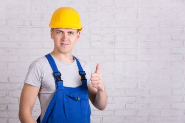 Mavi inşaatçı üniformalı genç adamın portresi beyaz tuğla duvarın üzerinde baş parmağıyla duruyor.