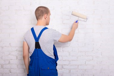 İş yerindeki genç bir ressamın arkadan görünüşü boya silindirli beyaz tuğla bir duvar boyuyor.