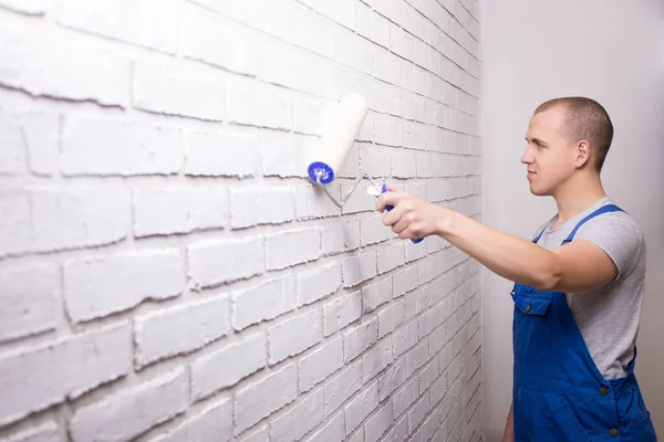 young man painter in uniform painting white brick wall with paint roller