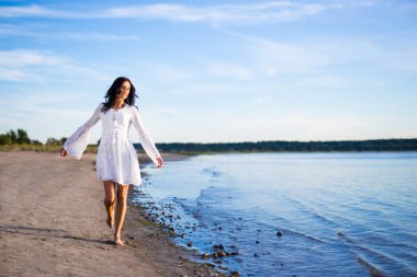 Kumsalda yürüyen beyaz elbiseli mutlu genç bir kadın.