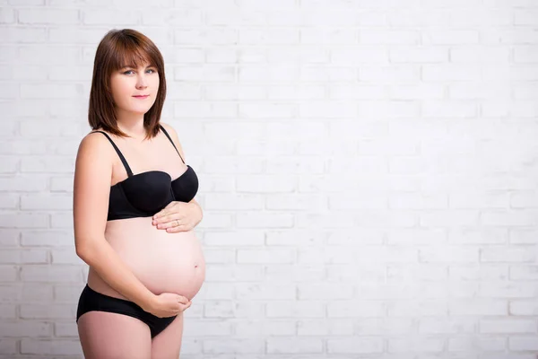 白いレンガの壁の背景に彼女の腹に触れるランジェリーの美しい妊婦 Copyspace — ストック写真