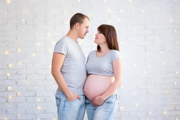 幸せな妊娠中のカップルの肖像画クリスマスライトと白いレンガの壁の上にお互いを見て — ストック写真