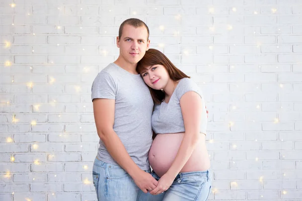 Portrait Happy Pregnant Couple Embracing White Brick Wall Christmas Lights — Stock Photo, Image