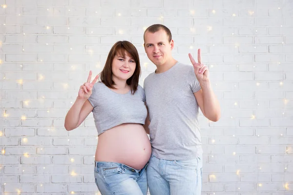 白いレンガの壁の上に勝利のサインを示すかわいい妊娠中のカップル — ストック写真